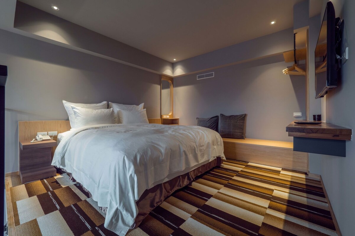 Una camera d'albergo moderna con un letto grande, un comodino con il telefono, un piccolo tavolino da notte con una lampada, una TV a parete e un tappeto a righe.