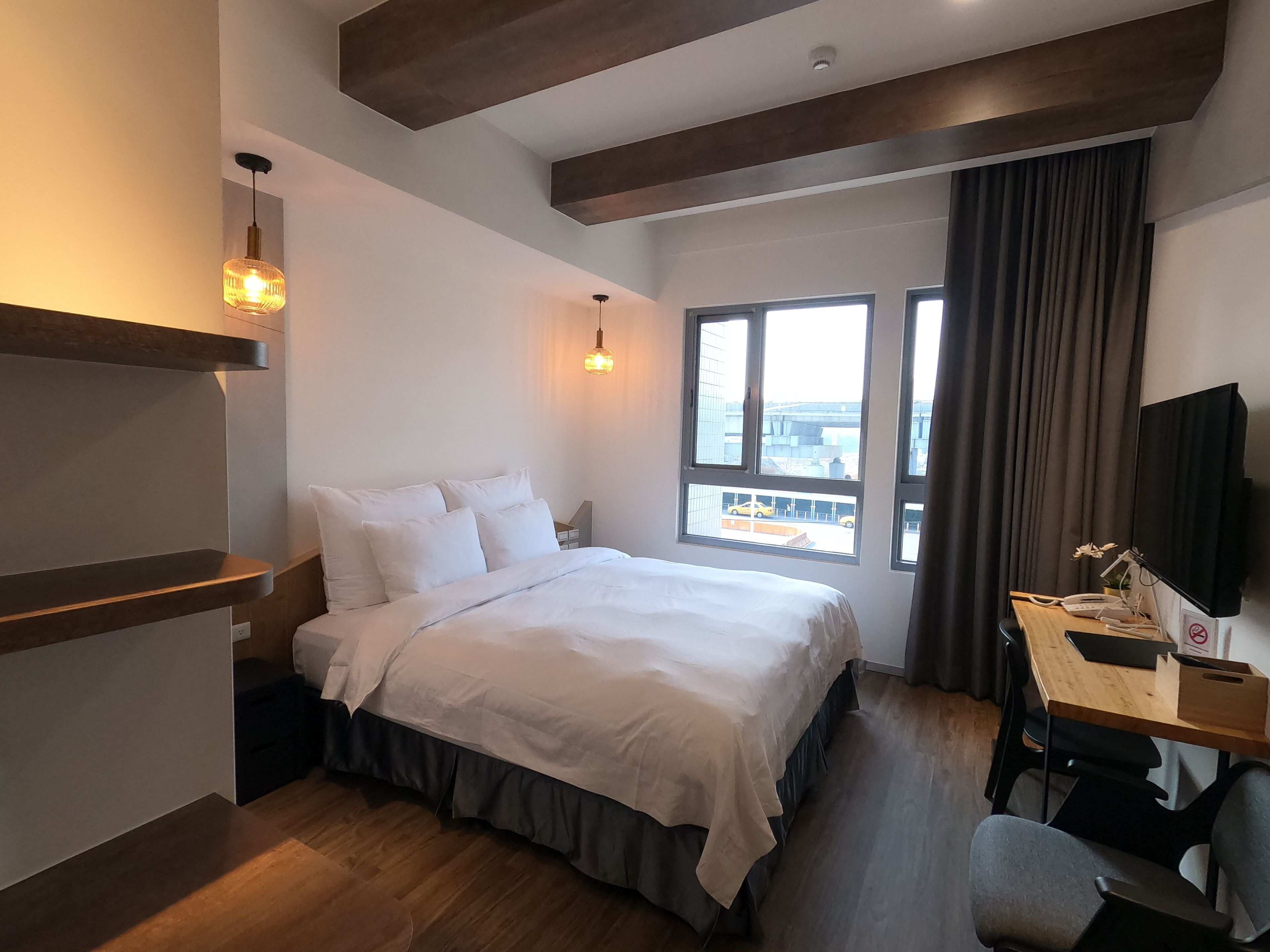 Una habitación de hotel moderna con una cama grande, un escritorio con una computadora y un televisor. Hay ventanas con vista a la ciudad y un balcón.