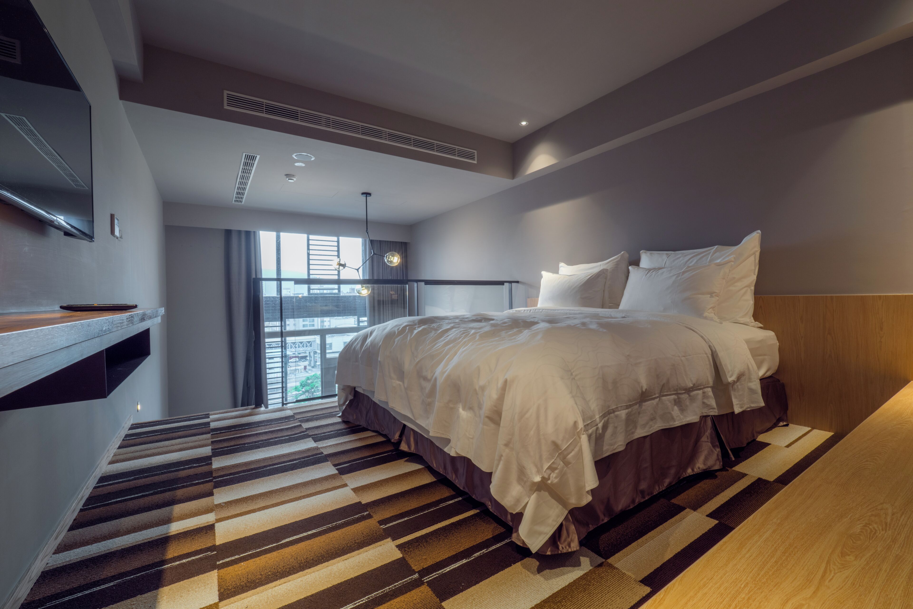 A modern hotel room with a large bed, a flat-screen TV, and a view of the city through the window.