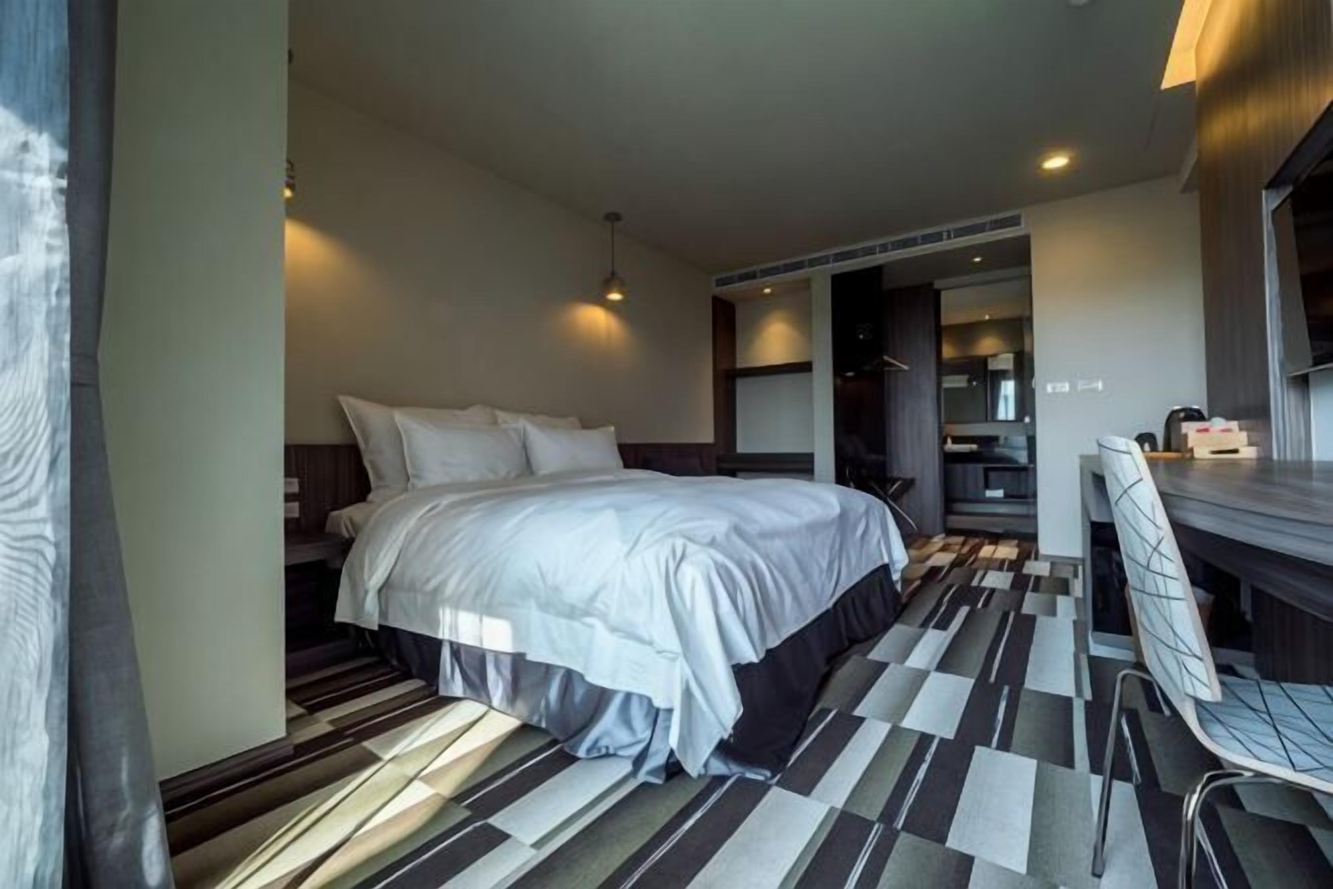 Camera d'albergo moderna con un letto grande, una scrivania e una sedia. Si intravedono una cucina e un bagno.