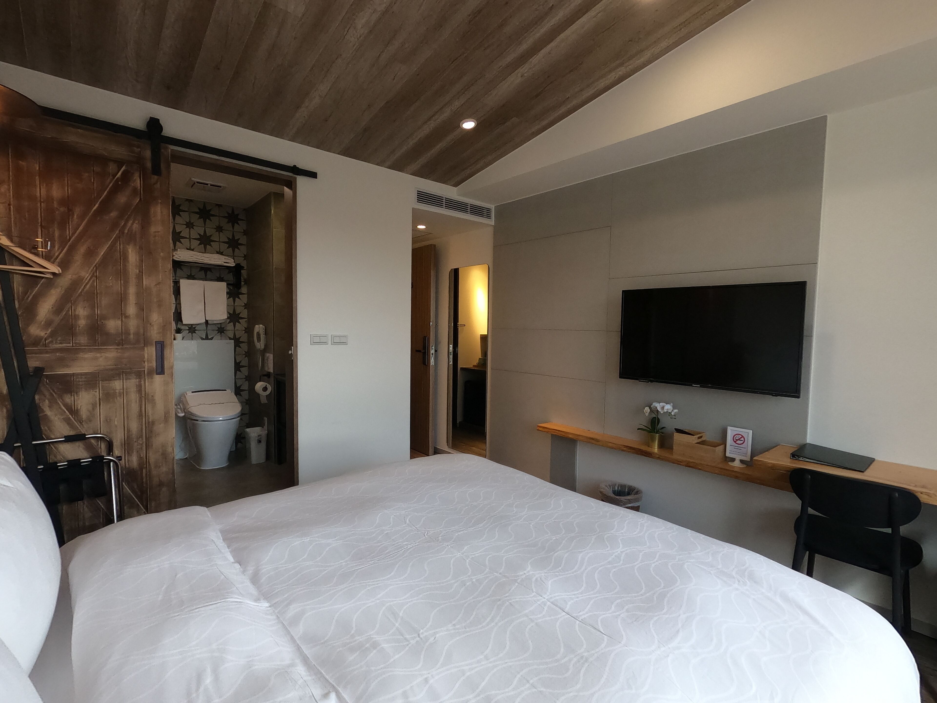 A bedroom with a large bed, a wooden ceiling, a TV mounted on the wall, and a small desk with a chair.