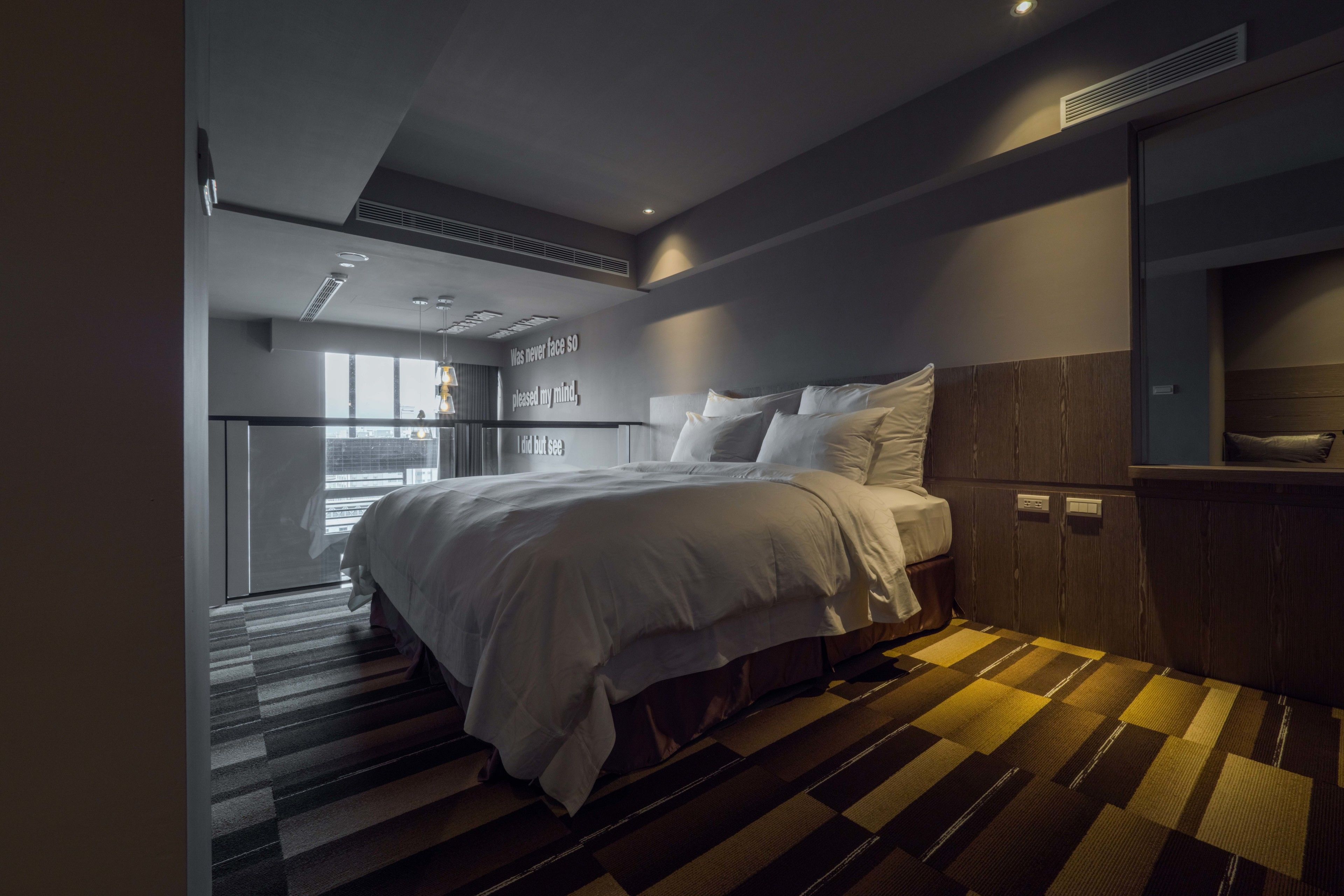 Una habitación de hotel moderna con una cama grande, cabecera de madera, escritorio integrado y vista a la ciudad a través de amplios ventanales.