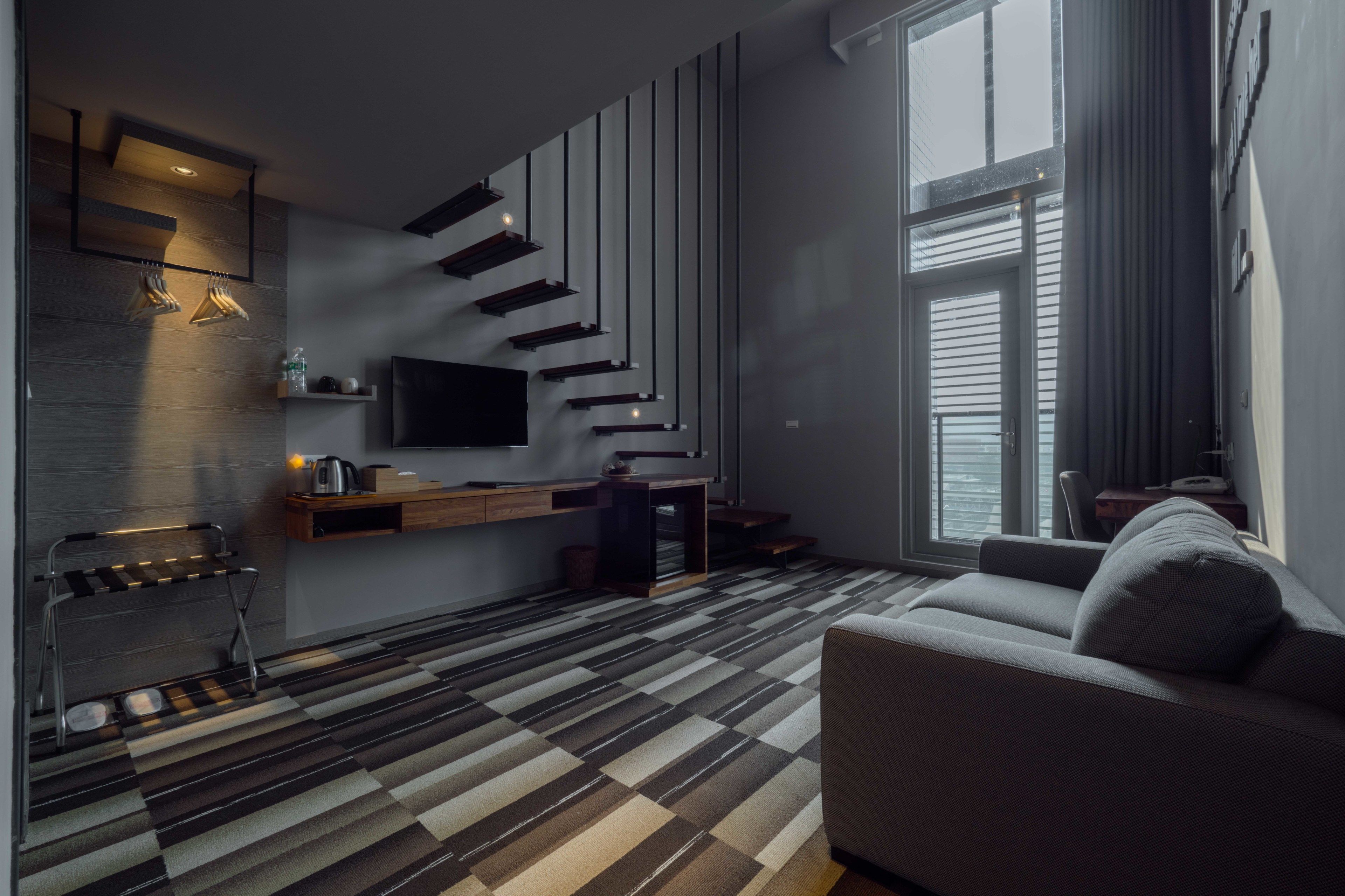 A modern living room with a sofa, a coffee table, a TV mounted on the wall, a staircase, and a window with blinds.