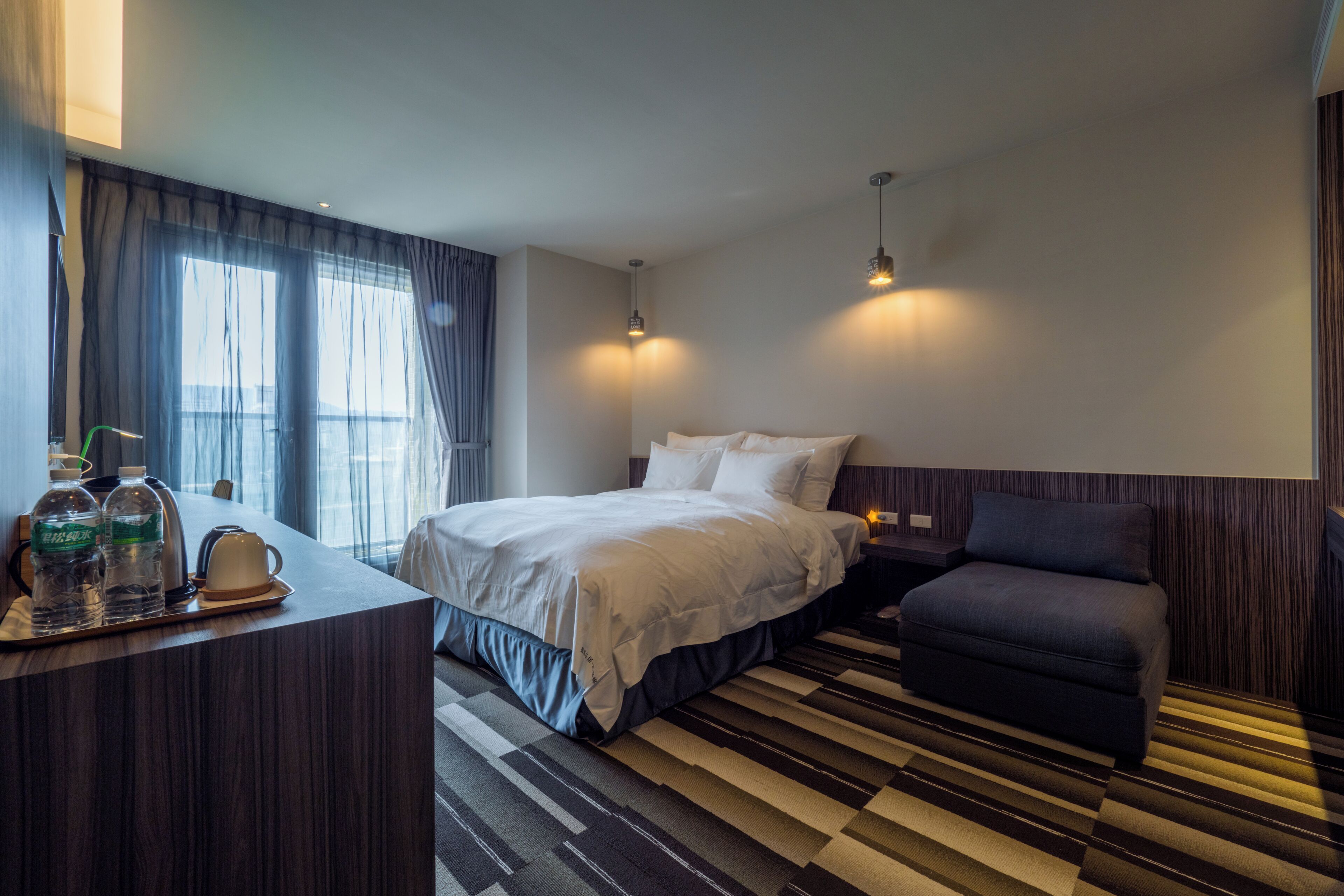 A hotel room with a bed, a sofa, a desk with water bottles, and a view of the outside through large windows.