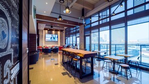Zona bar moderna con un lungo tavolo in legno, sgabelli da bar e ampie finestre con vista sulla città.