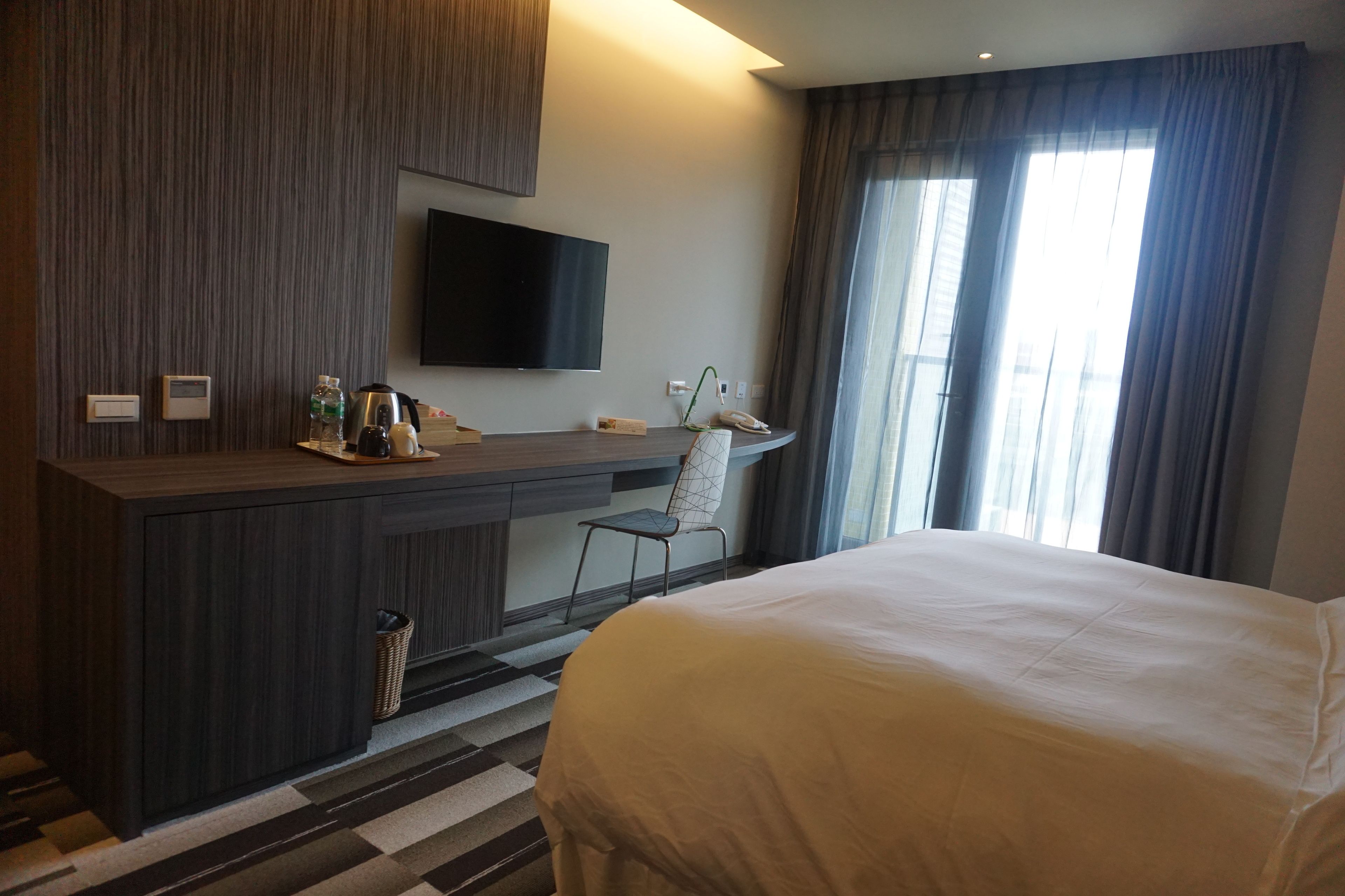 Una habitación de hotel moderna con una cama grande, un televisor de pantalla plana montado en la pared, un escritorio con una silla y un ventanal con cortinas.