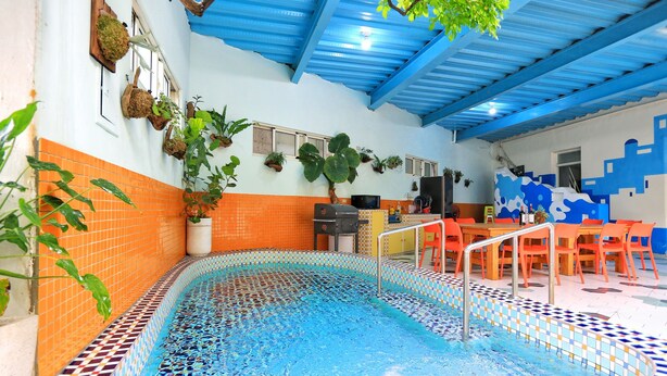 Una piscina interior con paredes y techo azulejados, rodeada de baldosas naranjas y blancas, y un comedor con sillas y mesas naranjas.