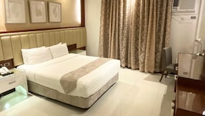 A hotel room with a large bed, a bedside table with a lamp, a chair, and a desk with a computer. - Crown Royale Hotel (Balanga)