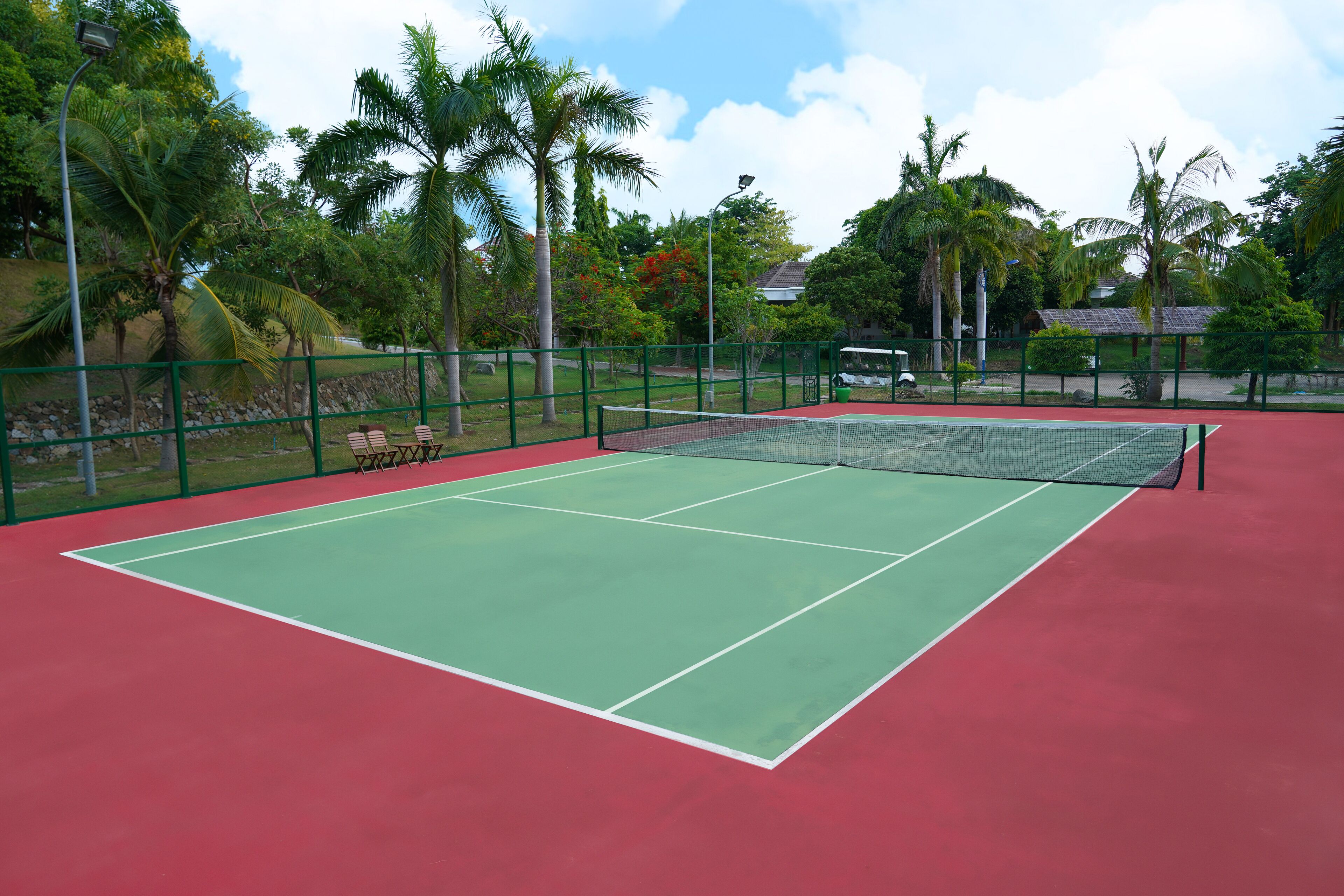 A tennis court with a green playing surface and a red surrounding area, surrounded by a green fence and palm trees.