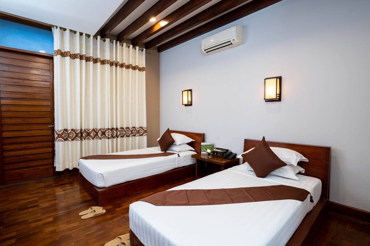 Quarto de hotel com duas camas, soalho em madeira, teto com vigas aparentes, aparelho de ar condicionado e janela com cortinas.