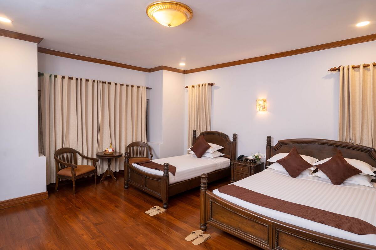 Quarto de hotel com duas camas de solteiro, uma cadeira de madeira, uma mesinha e uma mesa de cabeceira.