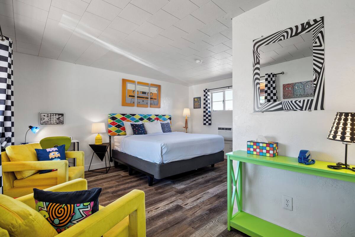 A modern bedroom with a bed, a green console table, a yellow armchair, and a large mirror.