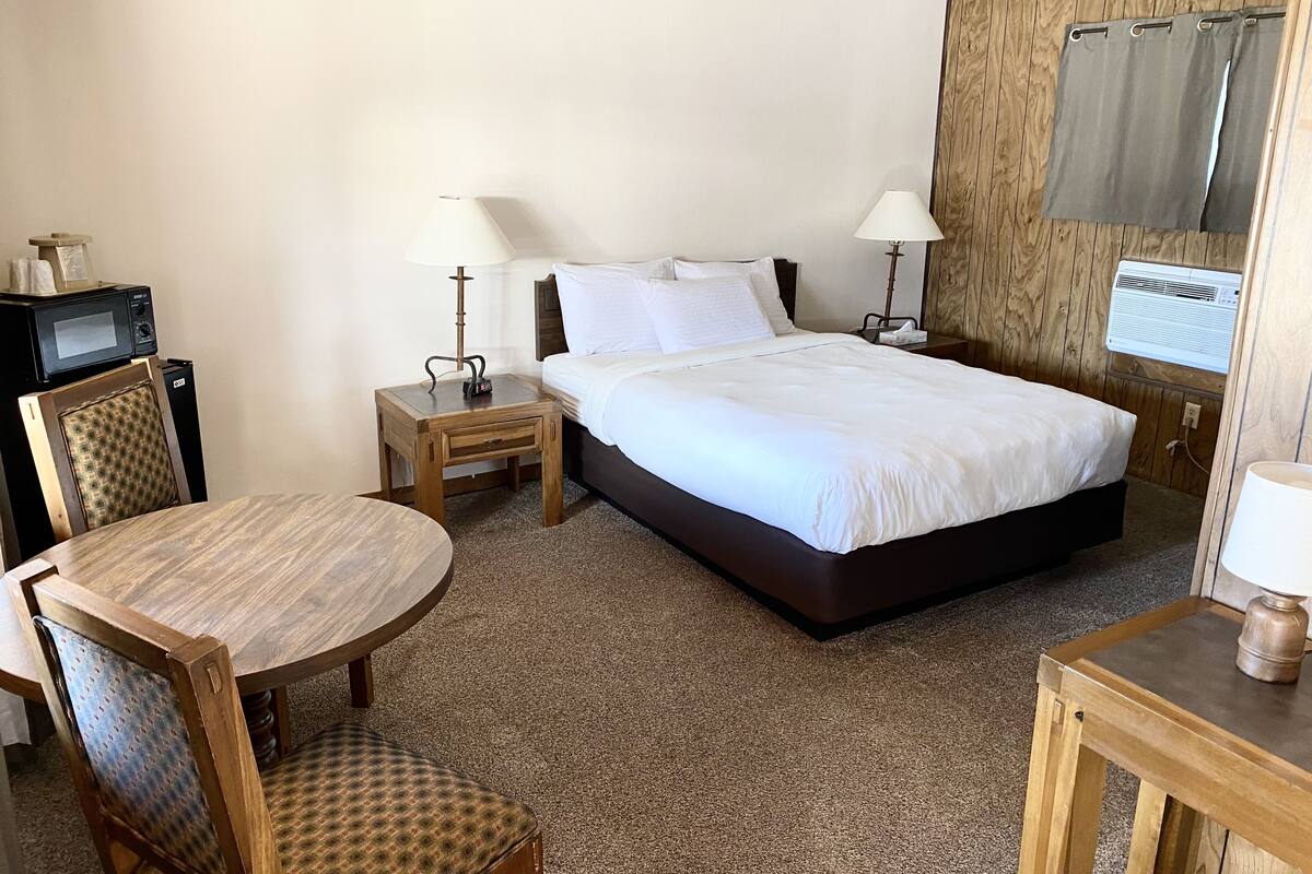 A hotel room with a bed, two bedside tables with lamps, a small desk with a chair, a microwave, and an air conditioning unit.