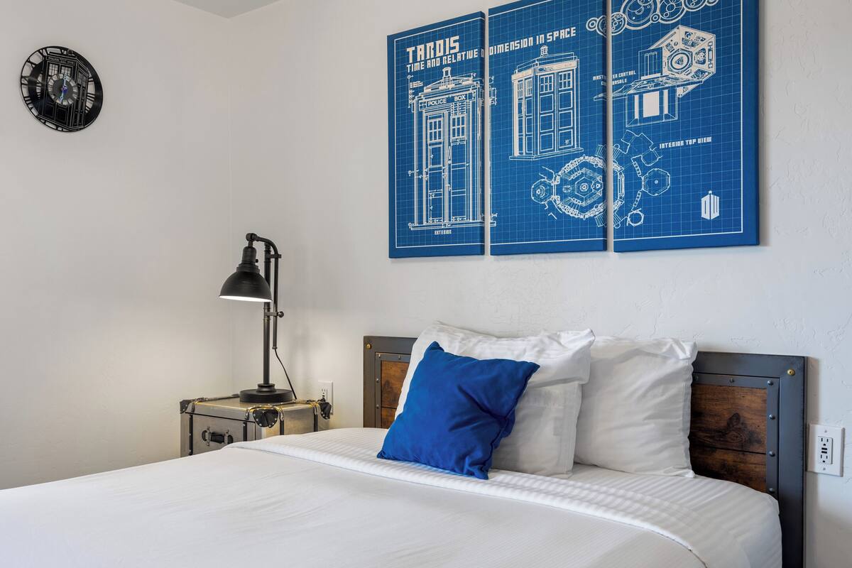 A bedroom with a bed, a nightstand, a lamp, a clock, and a blueprint artwork on the wall.