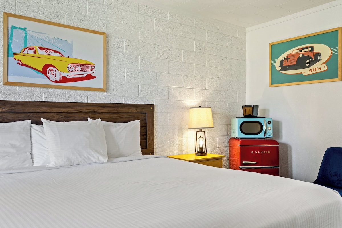 A hotel room with a bed, white bedding, a wooden headboard, a nightstand with a lamp, a vintage radio, and two framed pictures on the wall.