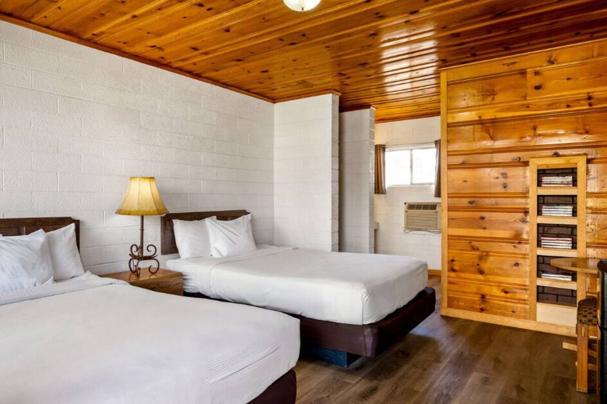 A room with two beds, a wooden ceiling, a brick wall, and a small table with a lamp.