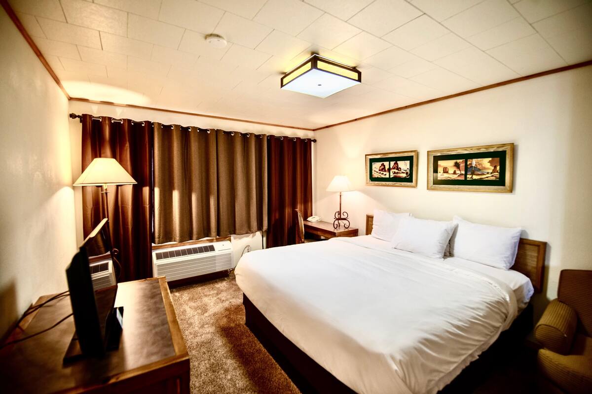 A hotel room with a large bed, two bedside lamps, a desk with a computer, a window with curtains, and two framed pictures on the wall.