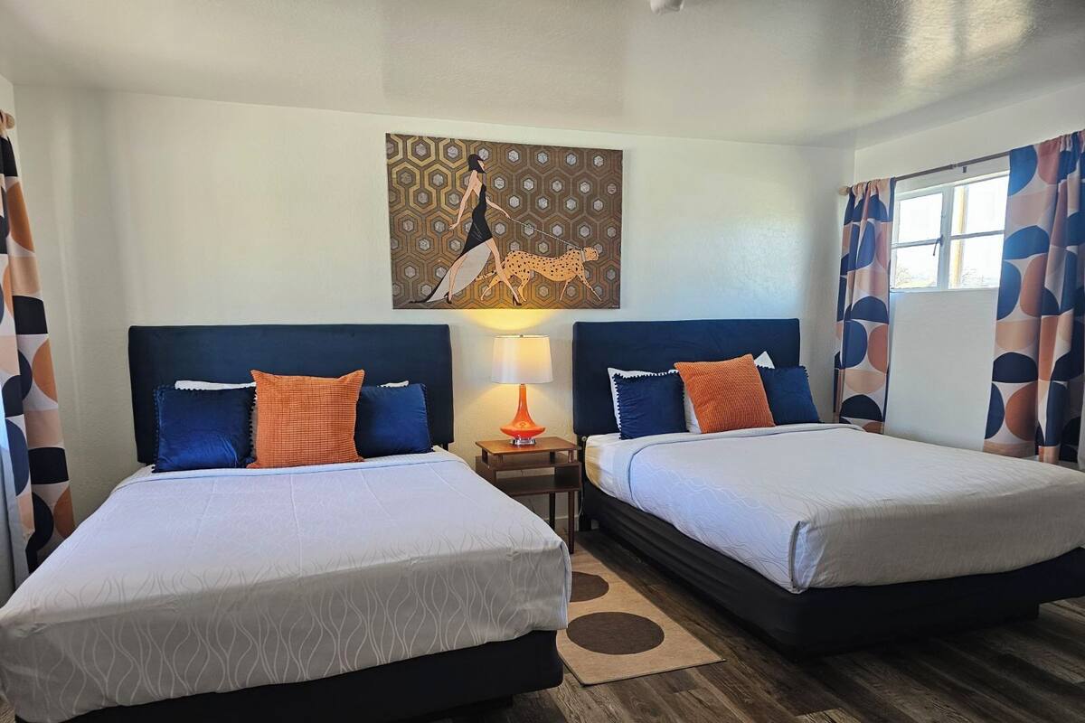 A hotel room with two beds, a nightstand, a lamp, a painting, and a window with curtains.