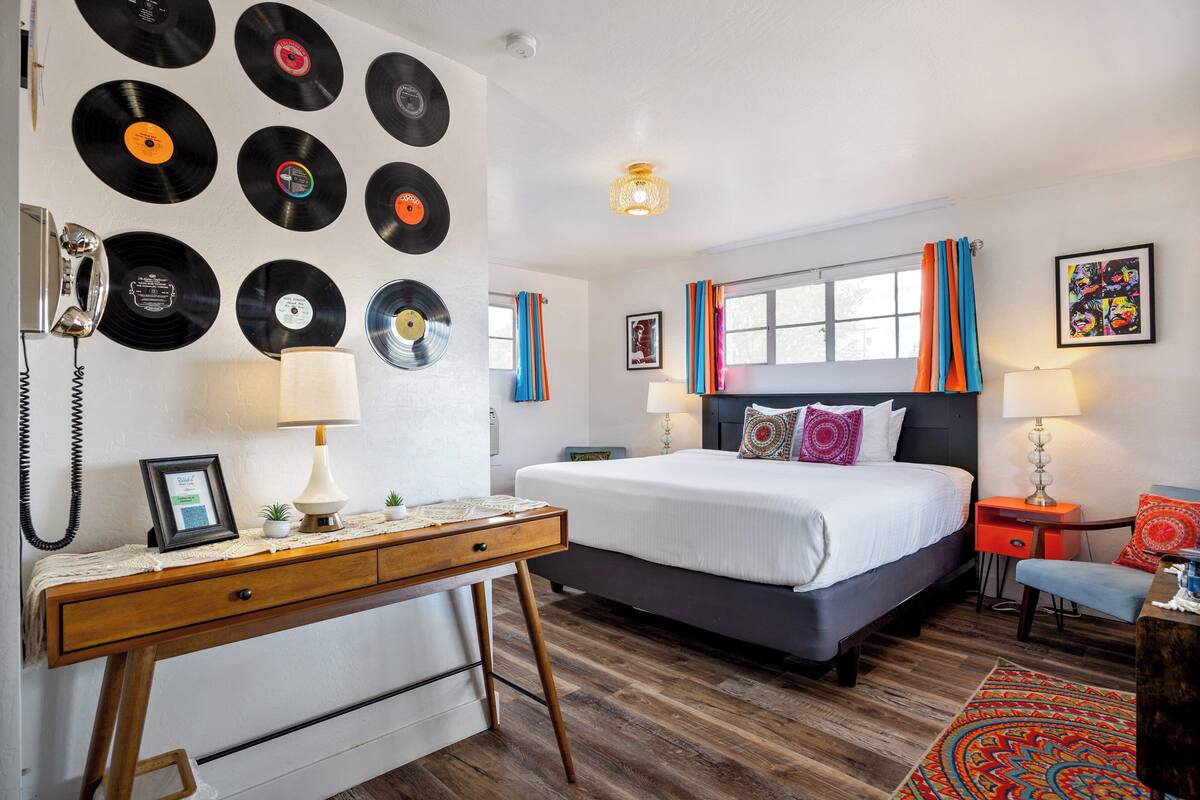 A bedroom with a bed, bedside tables, a desk, a wall of vinyl records, and a window with curtains.