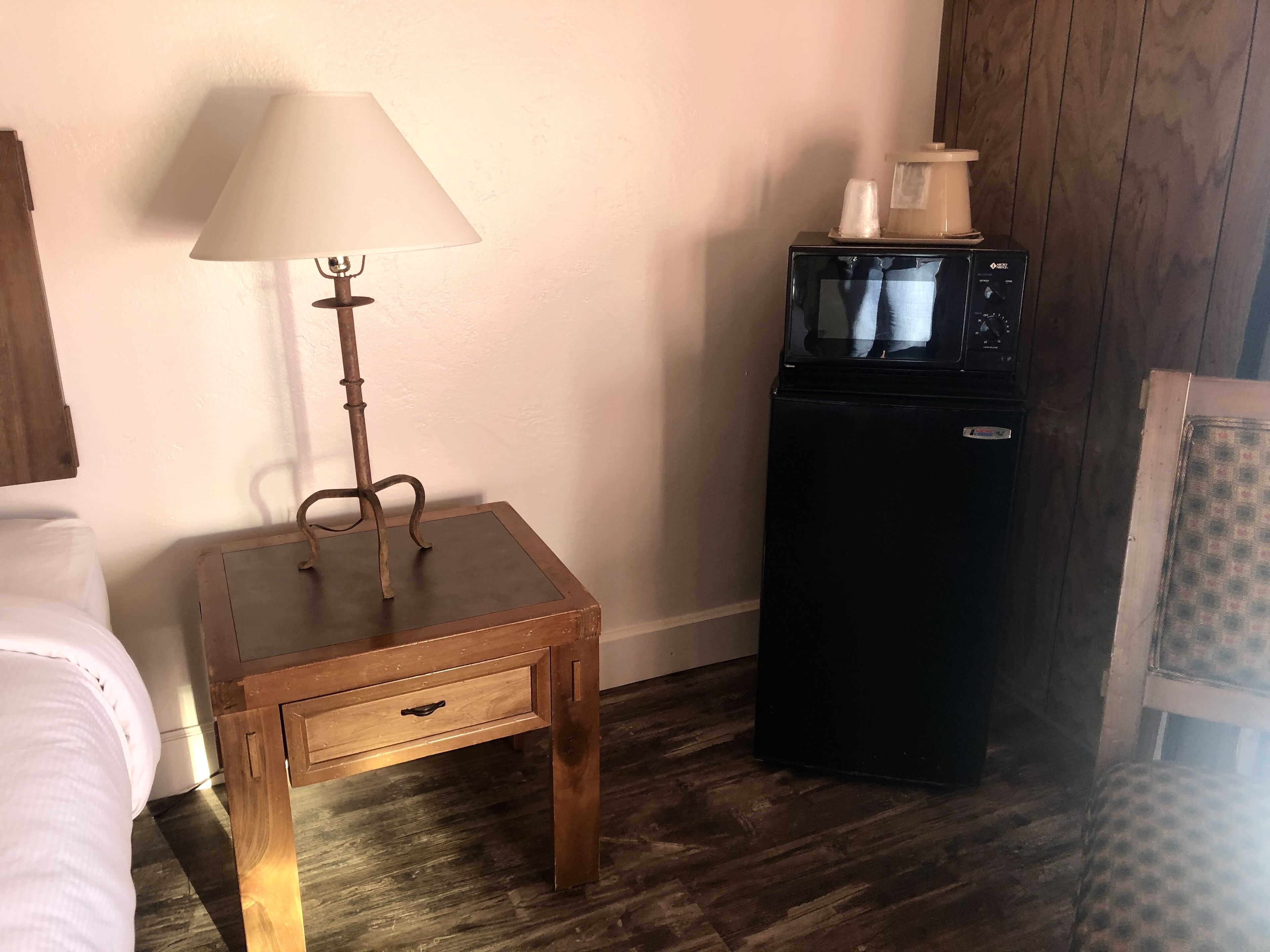 Una habitación pequeña con un mesita de noche de madera, una lámpara, un refrigerador negro y una cama blanca.