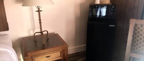 Una habitación pequeña con un mesita de noche de madera, una lámpara, un refrigerador negro y una cama blanca.