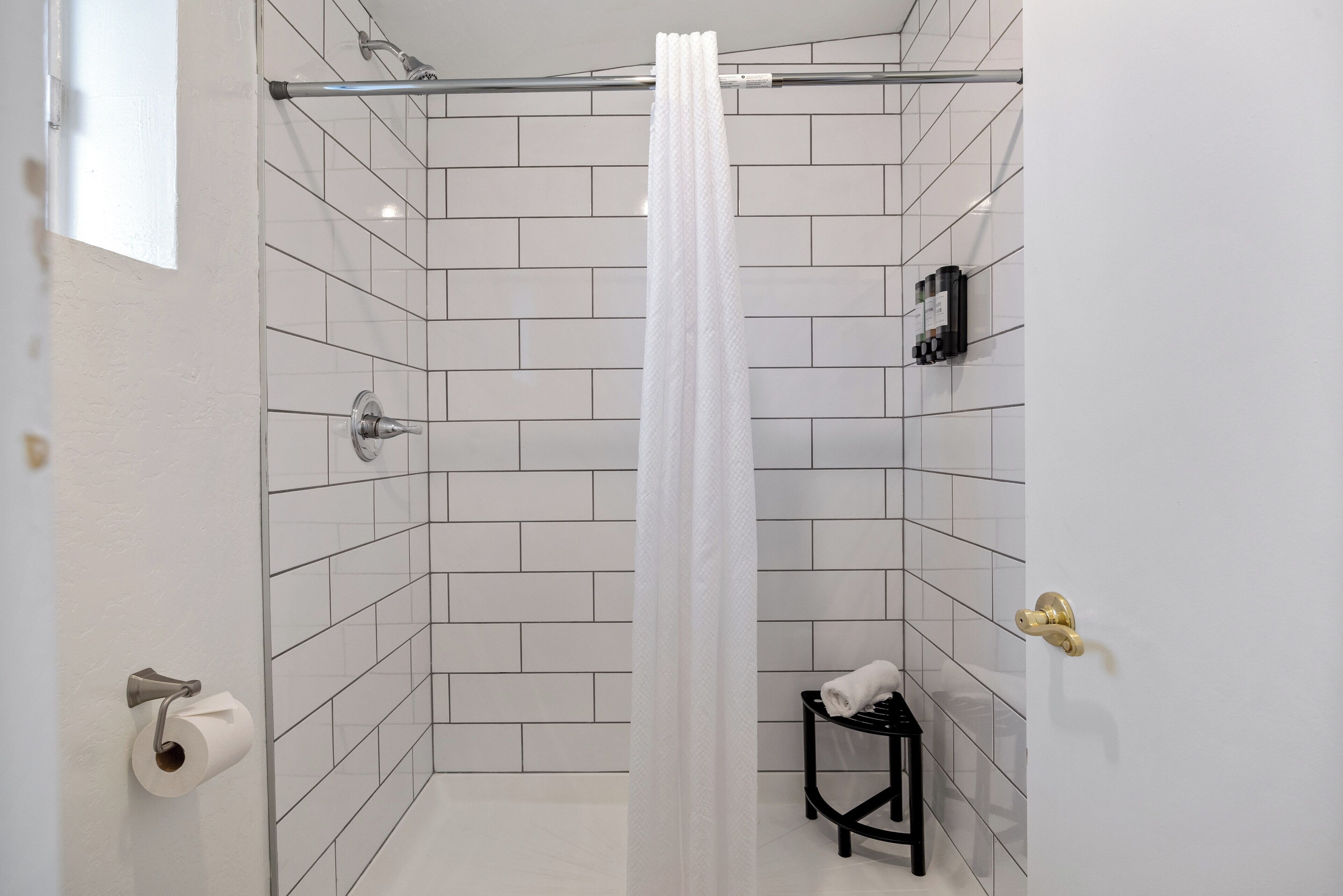 Un baño moderno con paredes de azulejos blancos, cortina de ducha, dispensador de papel higiénico y un pequeño taburete negro.