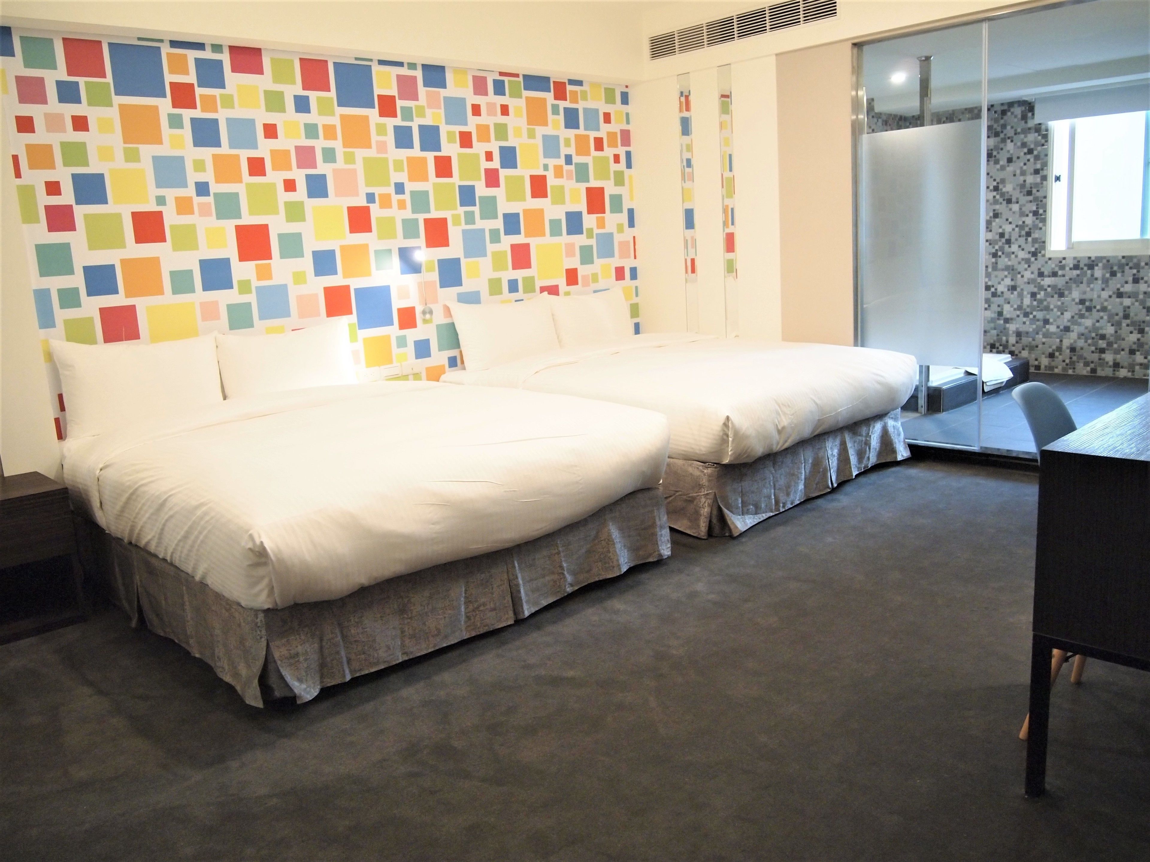 A hotel room with two beds, a desk, and a colorful mosaic wall.