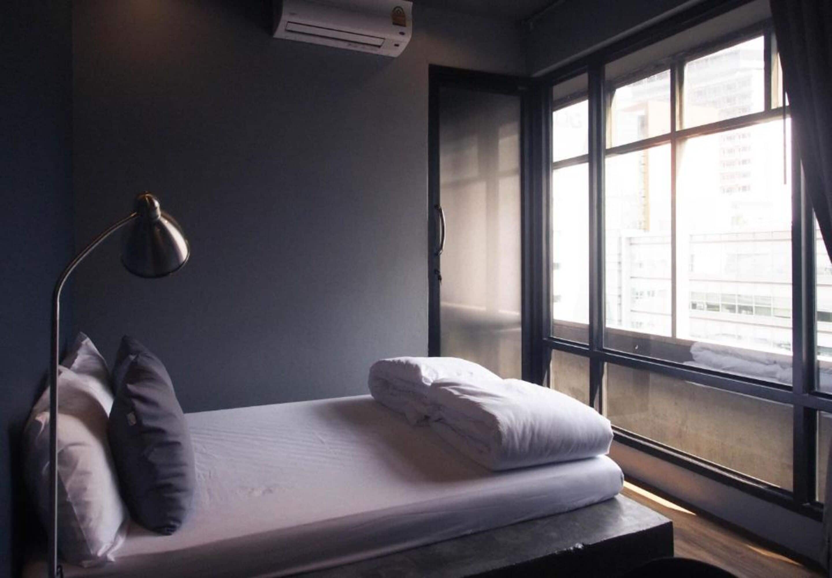 A single bed with white bedding, a desk with a lamp, and a window with a view of buildings.