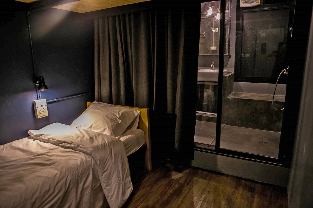 A small, dimly lit room with a single bed, a window with a view of a cityscape, and a bathroom with a sink and mirror.