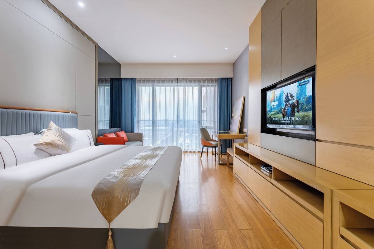 Moderni hotellihuone, jossa on suuri sänky, työpöytä, televisio ja suuret ikkunat, joista avautuu näkymä kaupunkiin.
