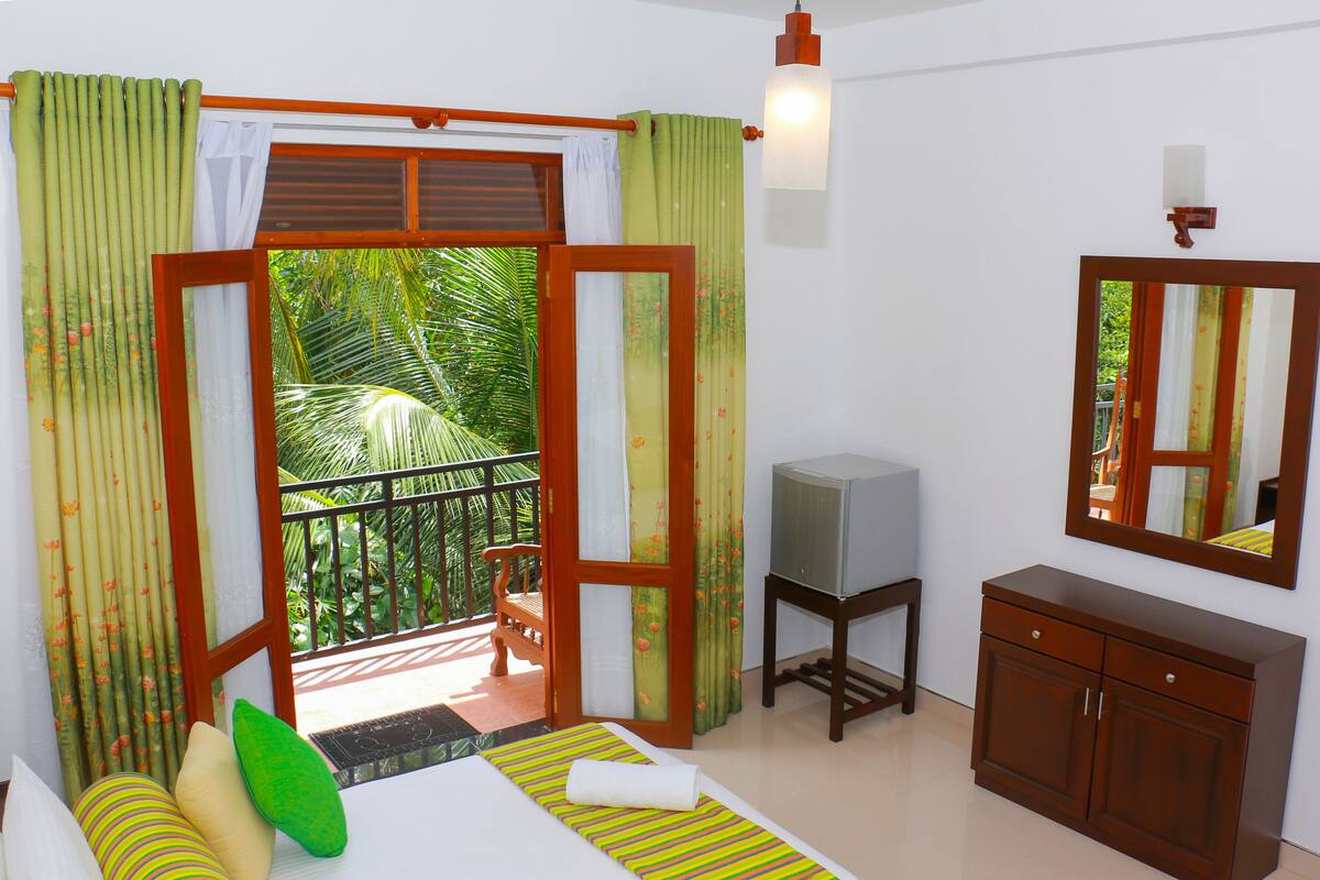 A bedroom with a bed, a TV, a wooden cabinet, and a balcony with a view of greenery.