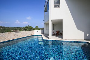 Una piscina con mosaicos, un edificio blanco con balcón y vista al paisaje.