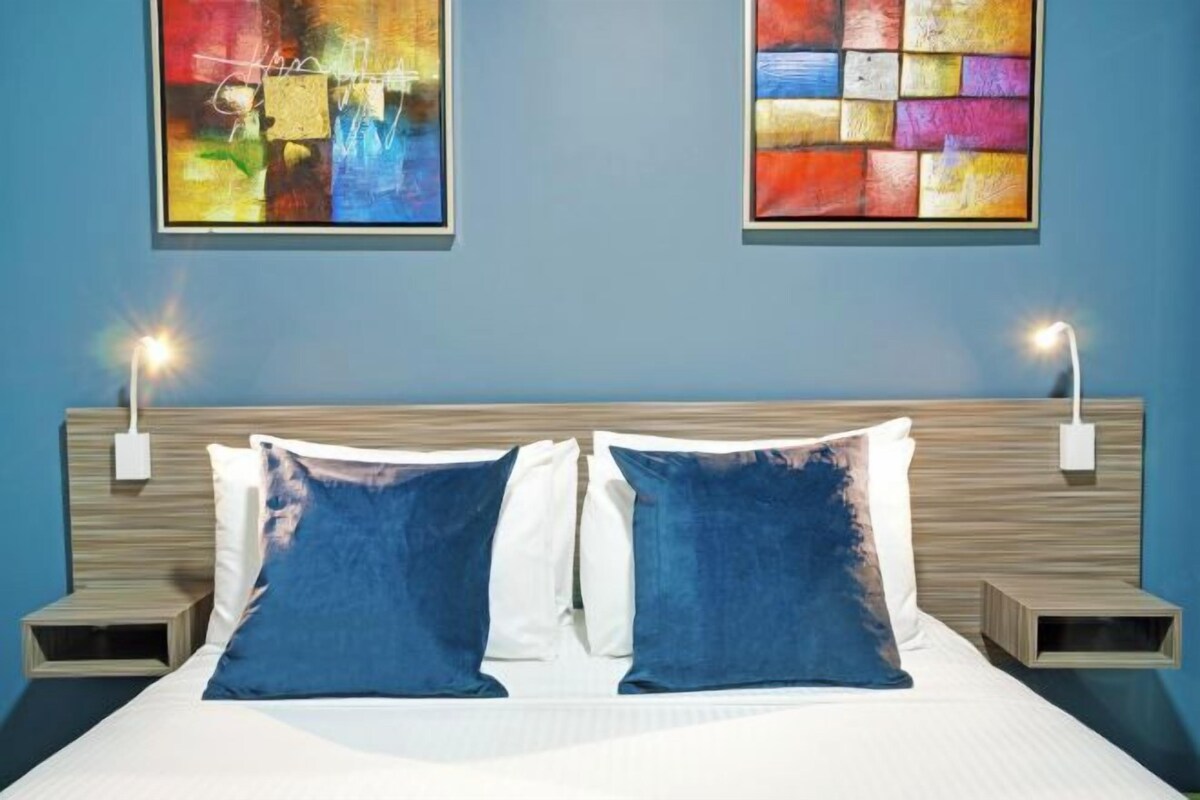 A hotel room with a bed, two wall-mounted nightstands, and two abstract paintings.