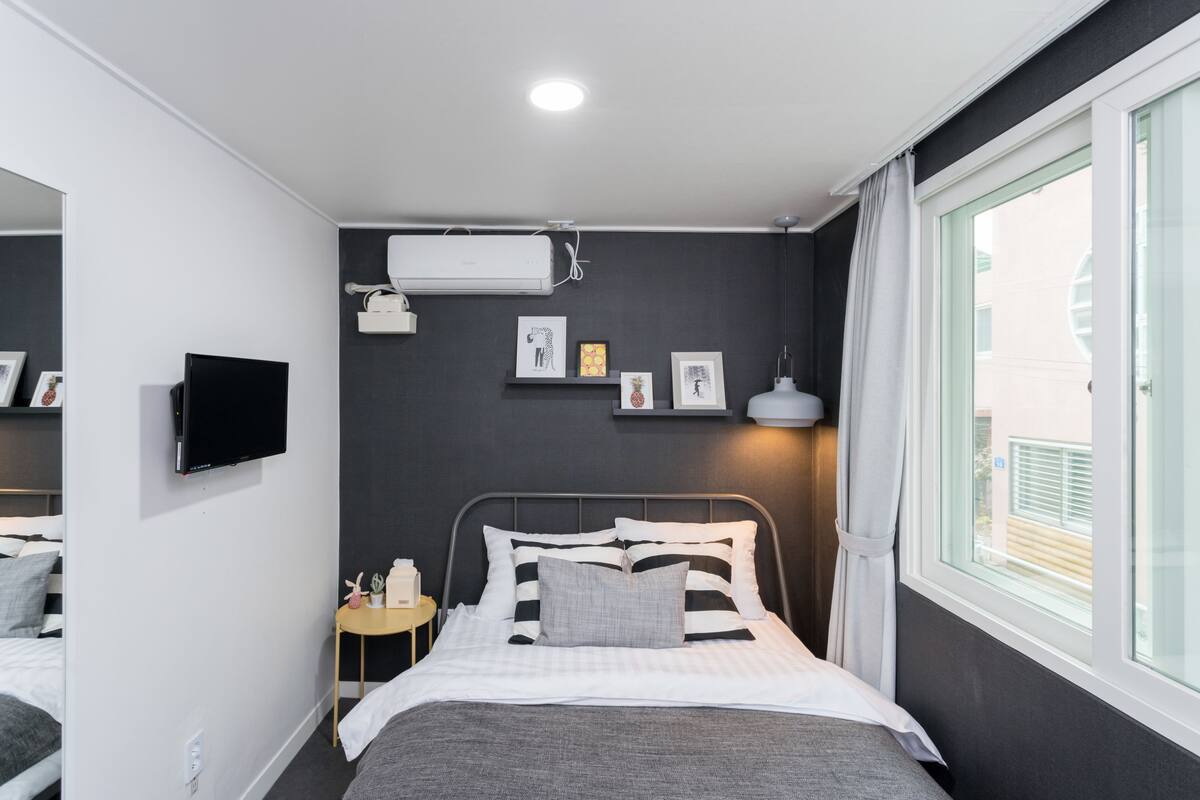 A modern hotel room with a bed, bedside table, wall-mounted TV, air conditioning unit, and a window with curtains.