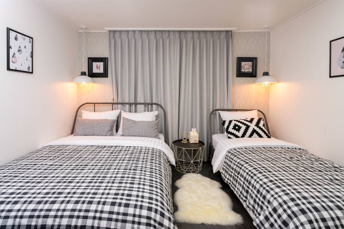 A hotel room with two beds, each with a checkered bedspread, a small round table with a lamp, and a white furry rug.