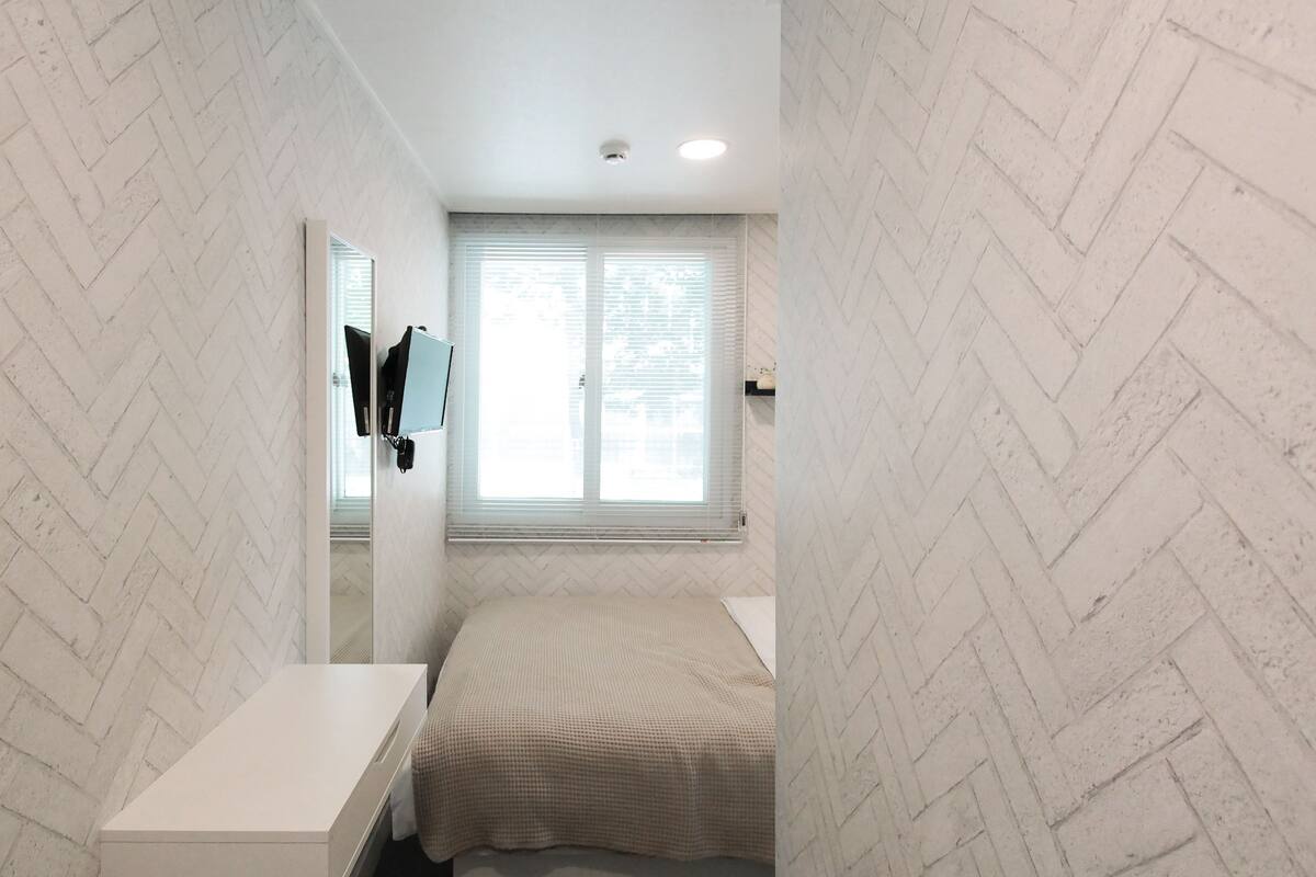 A bedroom with a bed, a bedside table, a mirror, and a window with blinds.