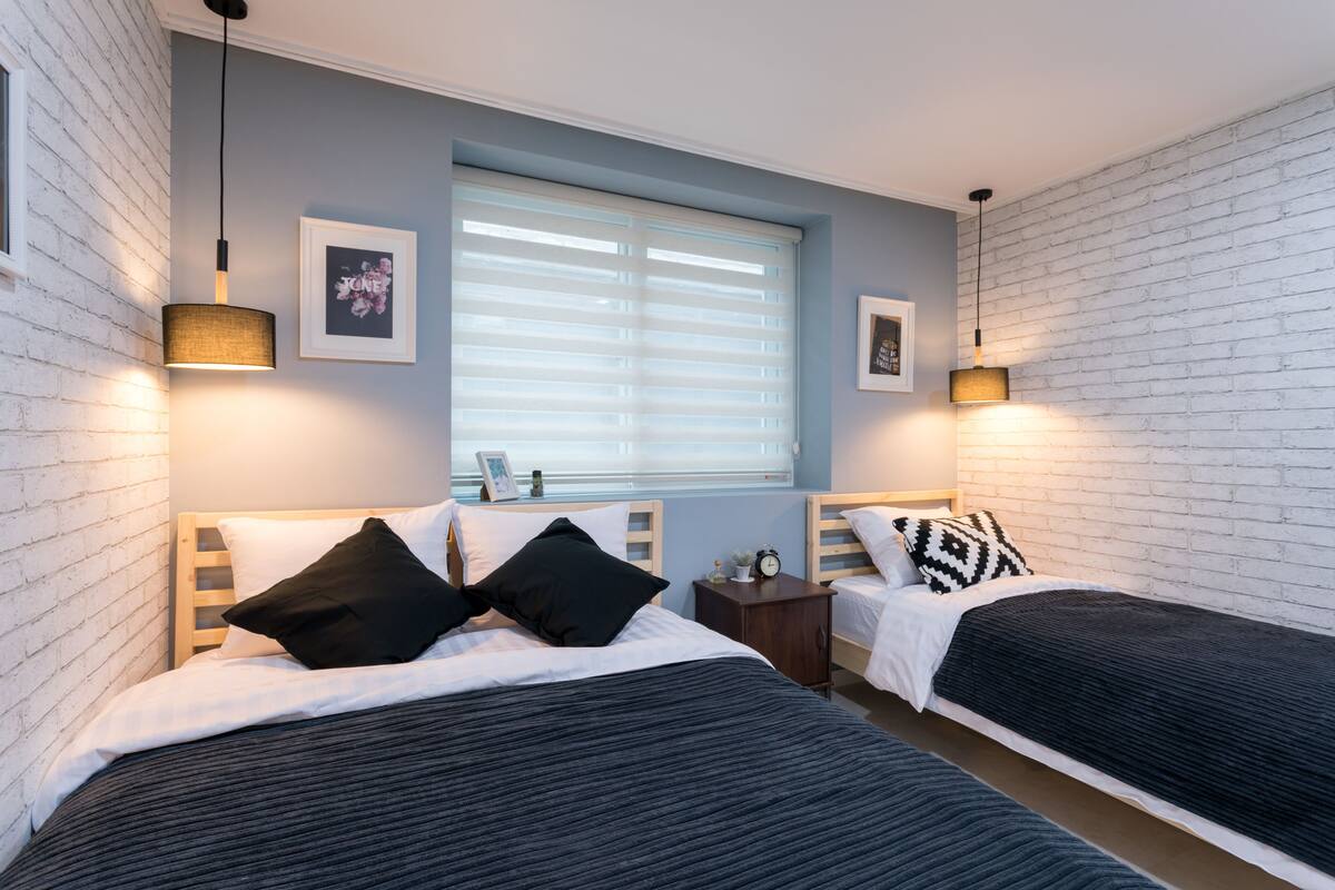 A bedroom with two beds, a window with blinds, and wall-mounted lamps.