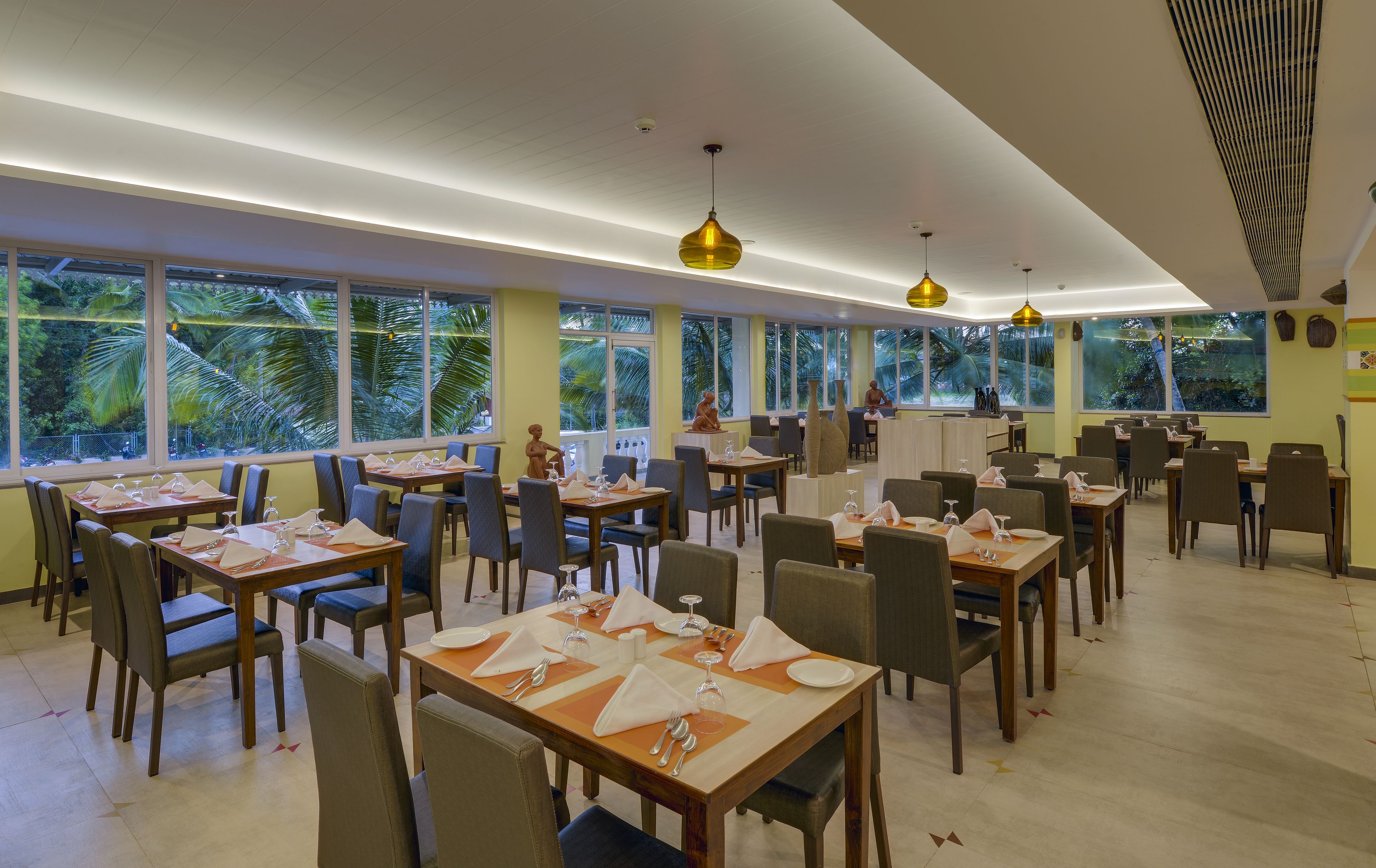 A restaurant with tables set for dining, large windows with a view of palm trees, and pendant lighting.