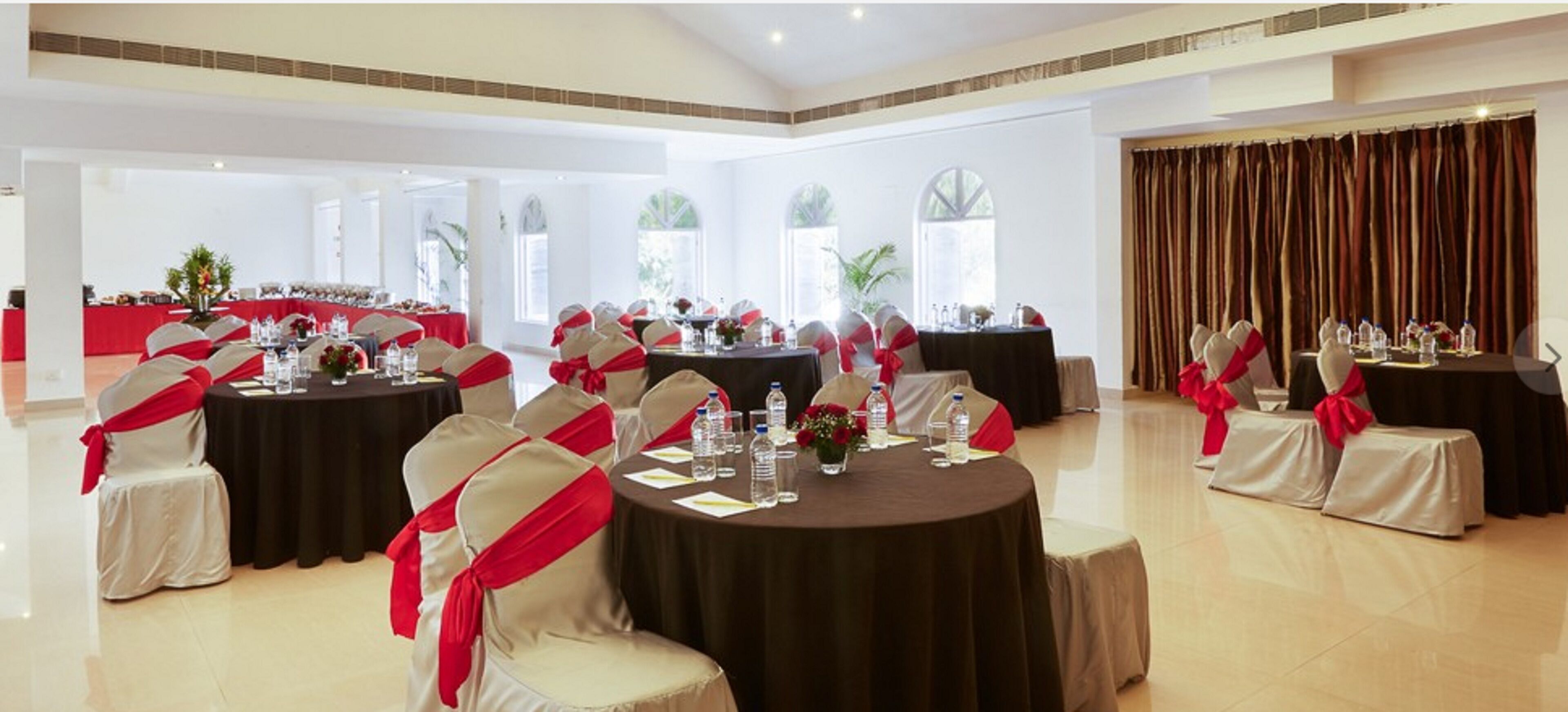Une salle de banquet avec des tables rondes, des chaises ornées de rubans rouges et une grande fenêtre avec des rideaux.