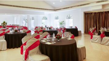 Une salle de banquet avec des tables rondes, des chaises ornées de rubans rouges et une grande fenêtre avec des rideaux.