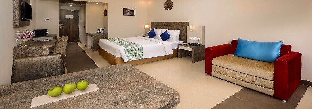 A hotel room with a bed, a red sofa, a dining table with apples, and a TV.