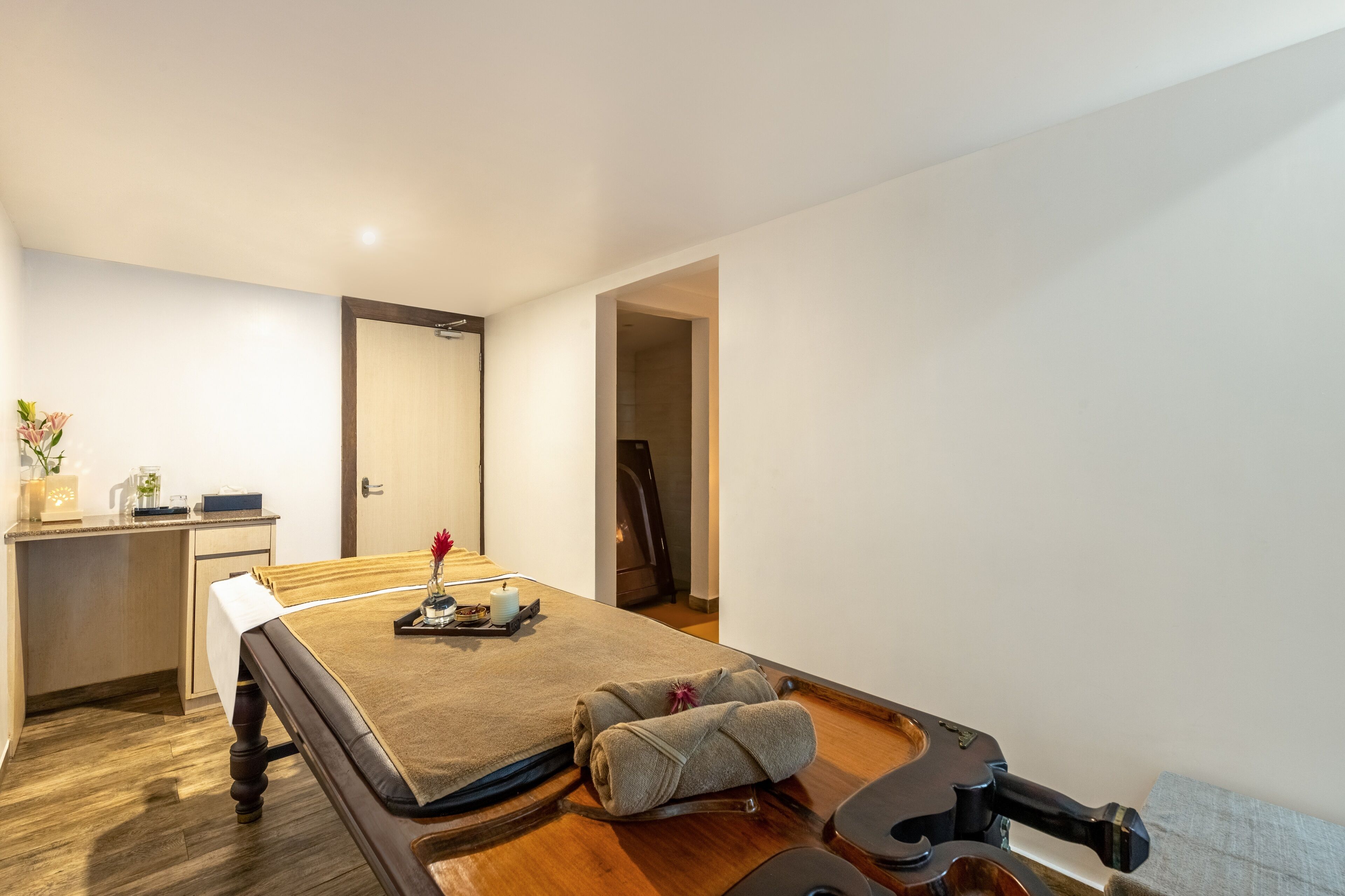 A modern hotel room with a wooden floor, a bed with a brown headboard, a desk with a lamp, and a small plant.