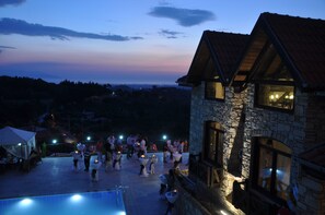 Um edifício de pedra com telhado em telhas, piscina e terraço com mesas e cadeiras.