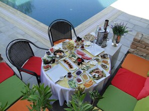 Una mesa redonda con mantel blanco, rodeada de sillas mullidas y coloridas, ubicada al aire libre con una piscina al fondo.