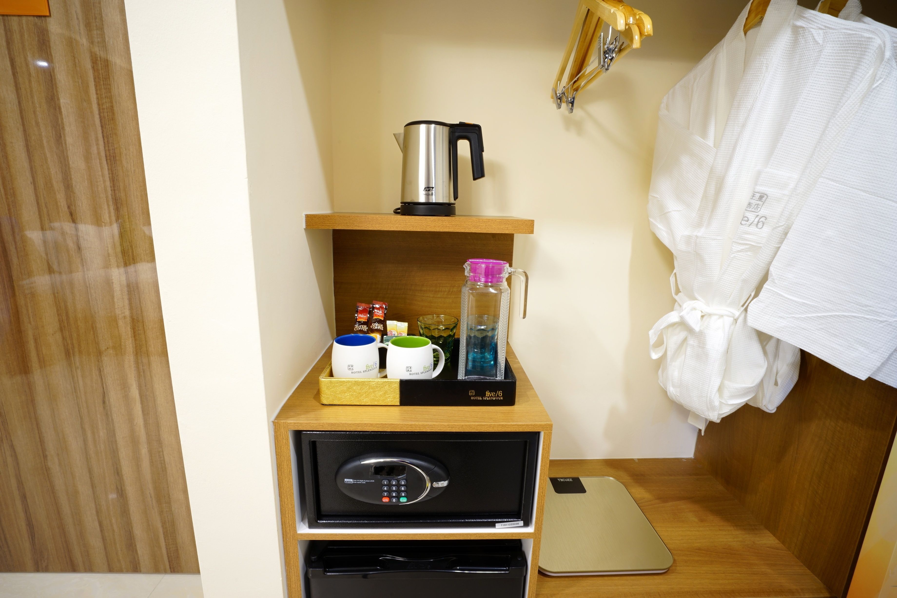 Habitación de hotel con repisa de madera que sostiene una tetera, una jarrón de vidrio y una bandeja con tazas y condimentos. Debajo de la repisa, hay un minibar negro con un teléfono y un refrigerador pequeño.