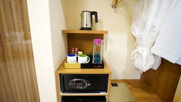 Habitación de hotel con repisa de madera que sostiene una tetera, una jarrón de vidrio y una bandeja con tazas y condimentos. Debajo de la repisa, hay un minibar negro con un teléfono y un refrigerador pequeño.