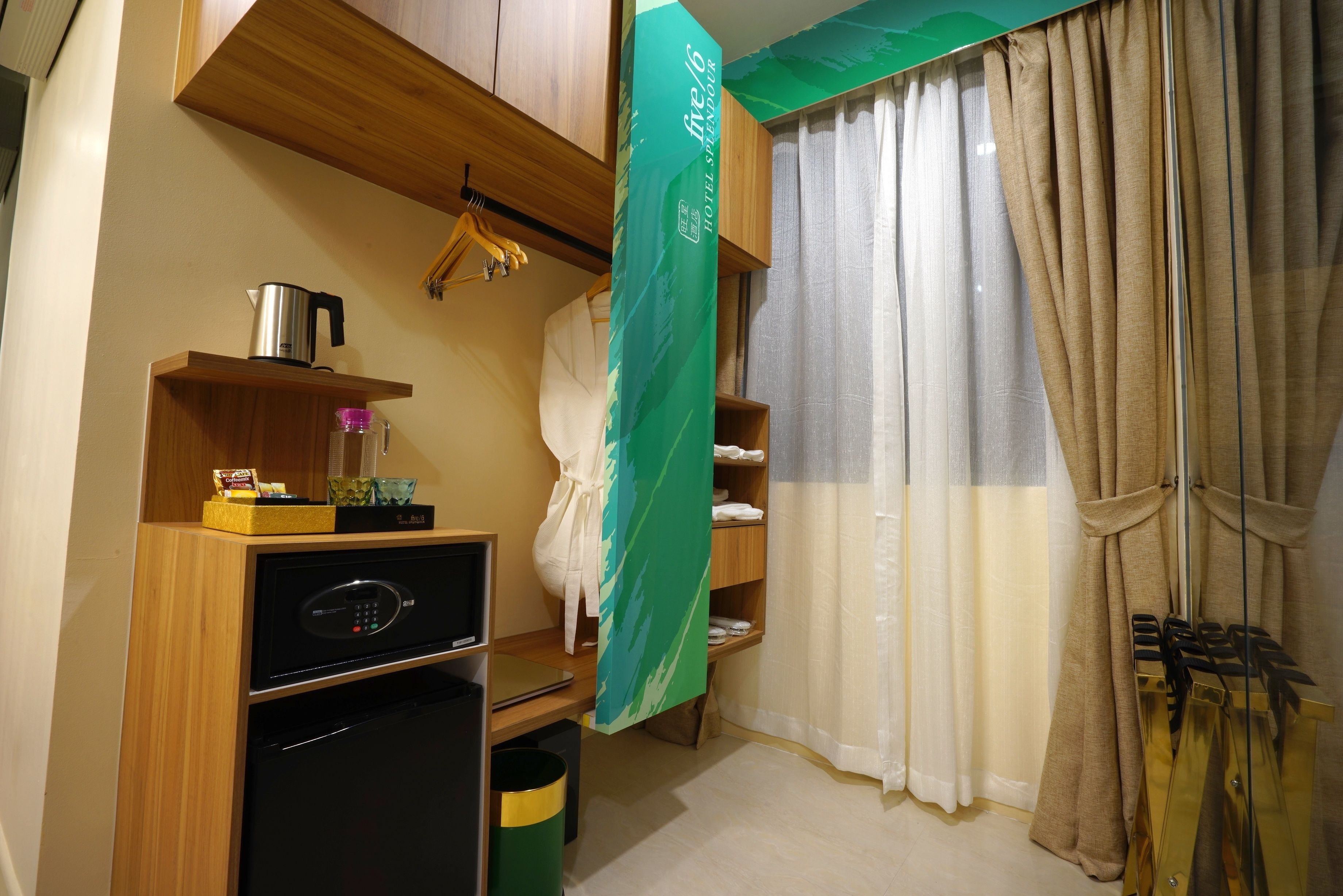 A compact hotel room with a kitchenette, including a microwave, a small refrigerator, and a coffee maker. There is a wooden shelf with a hanging garment and a towel rack. A green curtain partially covers a window with beige curtains.