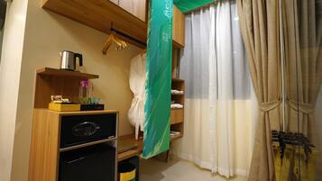 A compact hotel room with a kitchenette, including a microwave, a small refrigerator, and a coffee maker. There is a wooden shelf with a hanging garment and a towel rack. A green curtain partially covers a window with beige curtains.
