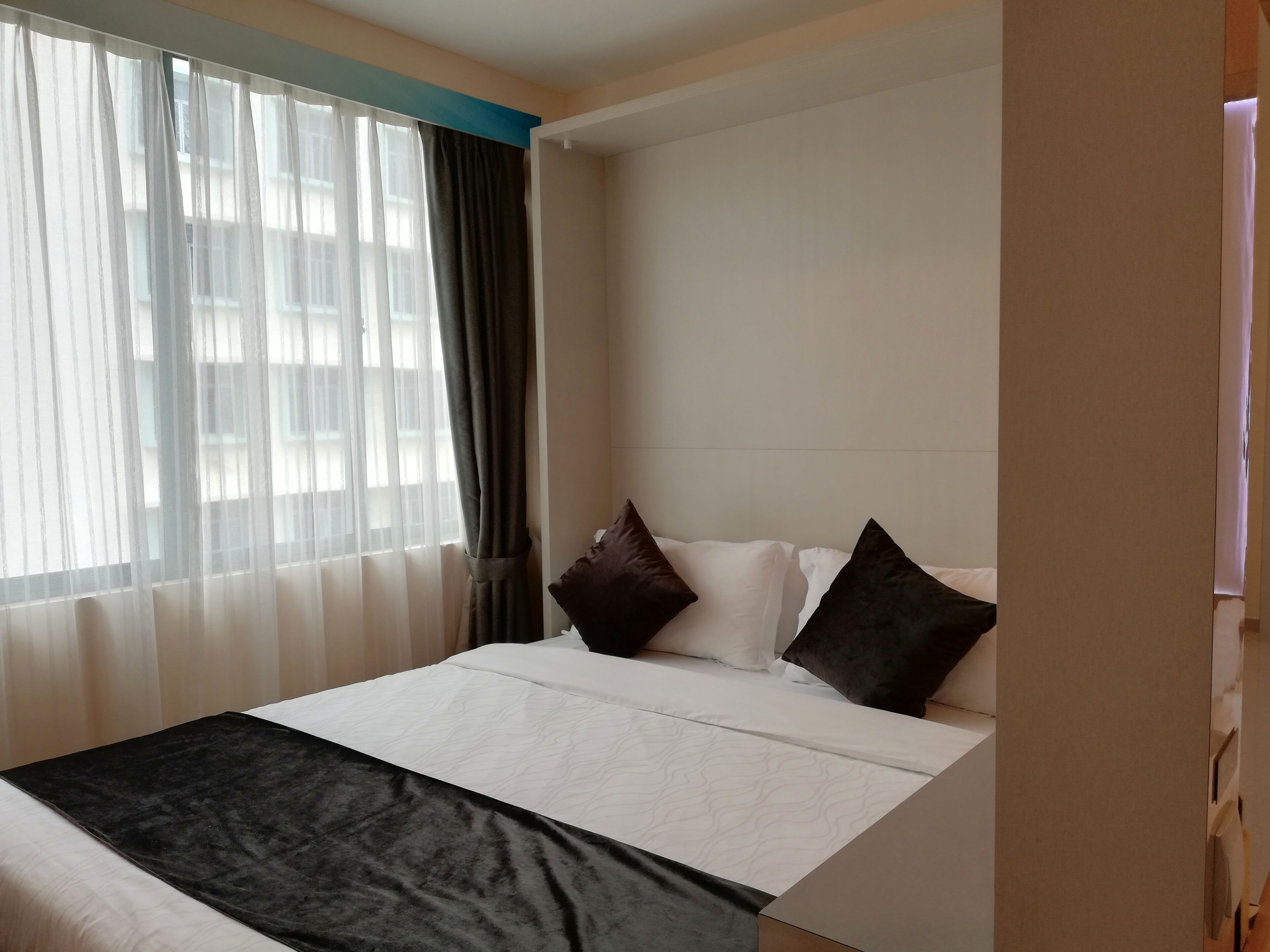 Habitación de hotel con una cama, ropa de cama blanca y negra, una ventana con cortinas traslúcidas y vistas a edificios.