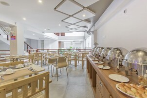 Ένας σύγχρονος χώρος εστίασης με πολλά τραπέζια και καρέκλες, μια σειρά από πάγκους με φαγητό σε στυλ μπουφέ, και μια σκάλα που οδηγεί σε ανώτερο επίπεδο.